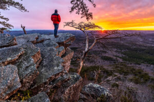 Man, person, standing on a rock high above a valley watching the sun setting in the distance