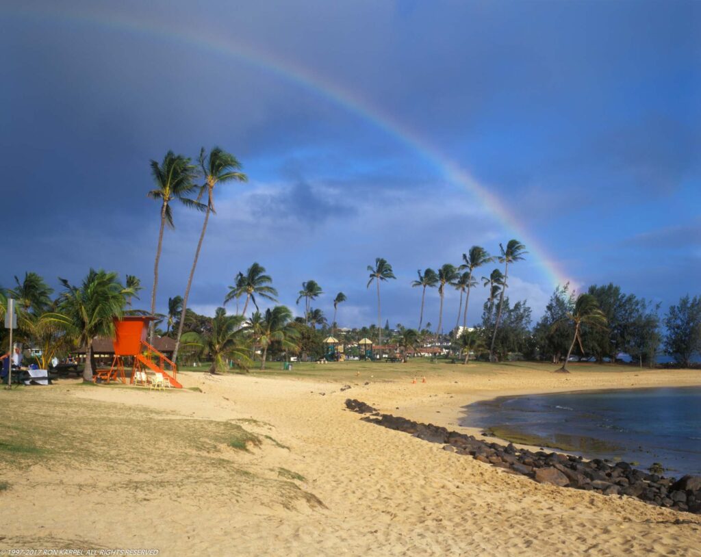 Poipu Beach and a rainbow. Note the old red lifeguard shade. The palm trees in the background are part of Poipu Beach Park which was voted #1 beach in America