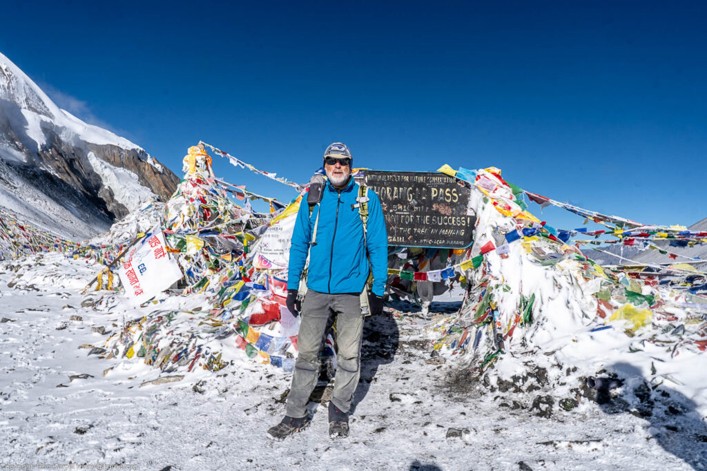 A person standing by a marker with a sign of Thorung La Pass with snow all around